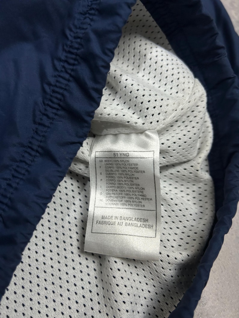 Op de afbeelding: Close-up van de binnenkant van een marineblauw kledingstuk, met een witte mesh voering. Een label toont tekst met materiaal samenstelling en "MADE IN BANGLADESH". De marineblauwe buitenkant van het kledingstuk is zichtbaar.