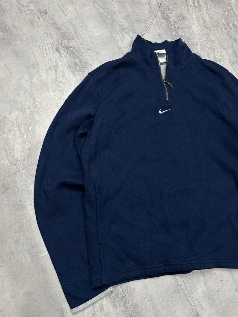 K&ouml;nnte beinhalten: Marineblaues Langarm-Pullover-Sweatshirt mit Viertelrei&szlig;verschluss und kleinem wei&szlig;em Nike-Logo auf der Brust. Der Kragen ist im Stehkragen-Stil. Das Sweatshirt hat eine lockere Passform.