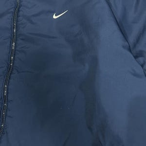 Op de afbeelding: Marineblauwe jas met een wit Nike swoosh-logo op de linkerborst. De jas heeft een volledige ritssluiting en een eenvoudig, strak ontwerp. Het materiaal lijkt lichtgewicht en geschikt om in lagen te dragen.