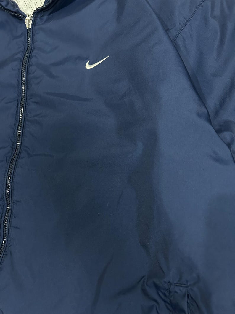 Op de afbeelding: Marineblauwe jas met een wit Nike swoosh-logo op de linkerborst. De jas heeft een volledige ritssluiting en een eenvoudig, strak ontwerp. Het materiaal lijkt lichtgewicht en geschikt om in lagen te dragen.