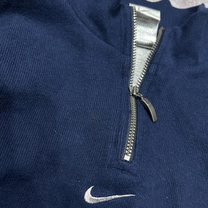 K&ouml;nnte beinhalten: Marineblaues Sweatshirt mit Viertelrei&szlig;verschluss, silbernem Rei&szlig;verschluss und kleinem wei&szlig;em Nike-Swoosh-Logo auf der Brust. Das Sweatshirt hat einen hohen Kragen.