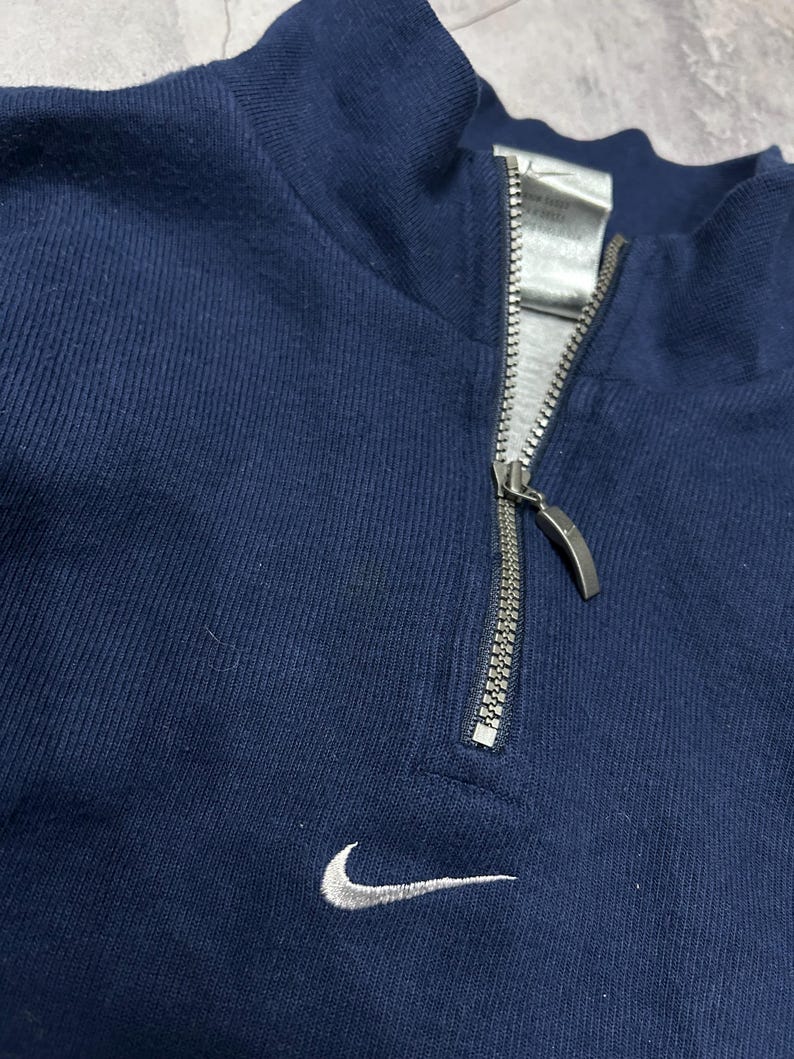 K&ouml;nnte beinhalten: Marineblaues Sweatshirt mit Viertelrei&szlig;verschluss, silbernem Rei&szlig;verschluss und kleinem wei&szlig;em Nike-Swoosh-Logo auf der Brust. Das Sweatshirt hat einen hohen Kragen.