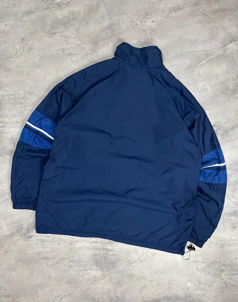 Op de afbeelding: Marineblau sportjack met een hoge kraag en een trekkoord aan de zoom. De jas heeft blauwe en witte strepen op de mouwen. De jas is gemaakt van een lichtgewicht materiaal.