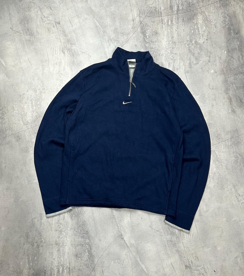 K&ouml;nnte beinhalten: Marineblaues Langarm-Pullover mit einem Viertel-Rei&szlig;verschluss und einem kleinen Nike-Logo auf der Brust. Der Kragen und die Manschetten haben einen hellgrauen Besatz. Das Oberteil ist aus weichem, bequemem Material.