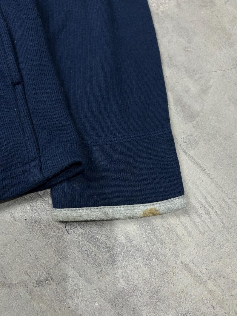 K&ouml;nnte beinhalten: Nahaufnahme eines marineblauen Sweatshirt-&Auml;rmels. Das B&uuml;ndchen hat einen grauen Besatz mit einem braunen Fleck. Der Stoff scheint ein dickes Strickmaterial zu sein.