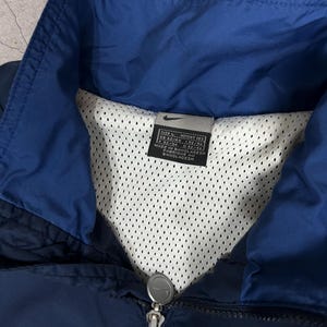Op de afbeelding: Marineblauwe en koningsblauwe Nike jas met mesh voering. De jas heeft een ritssluiting en een opstaande kraag. Het Nike-logo is zichtbaar op de ritstrekker. Het label geeft maat L aan en is gemaakt in Bangladesh.