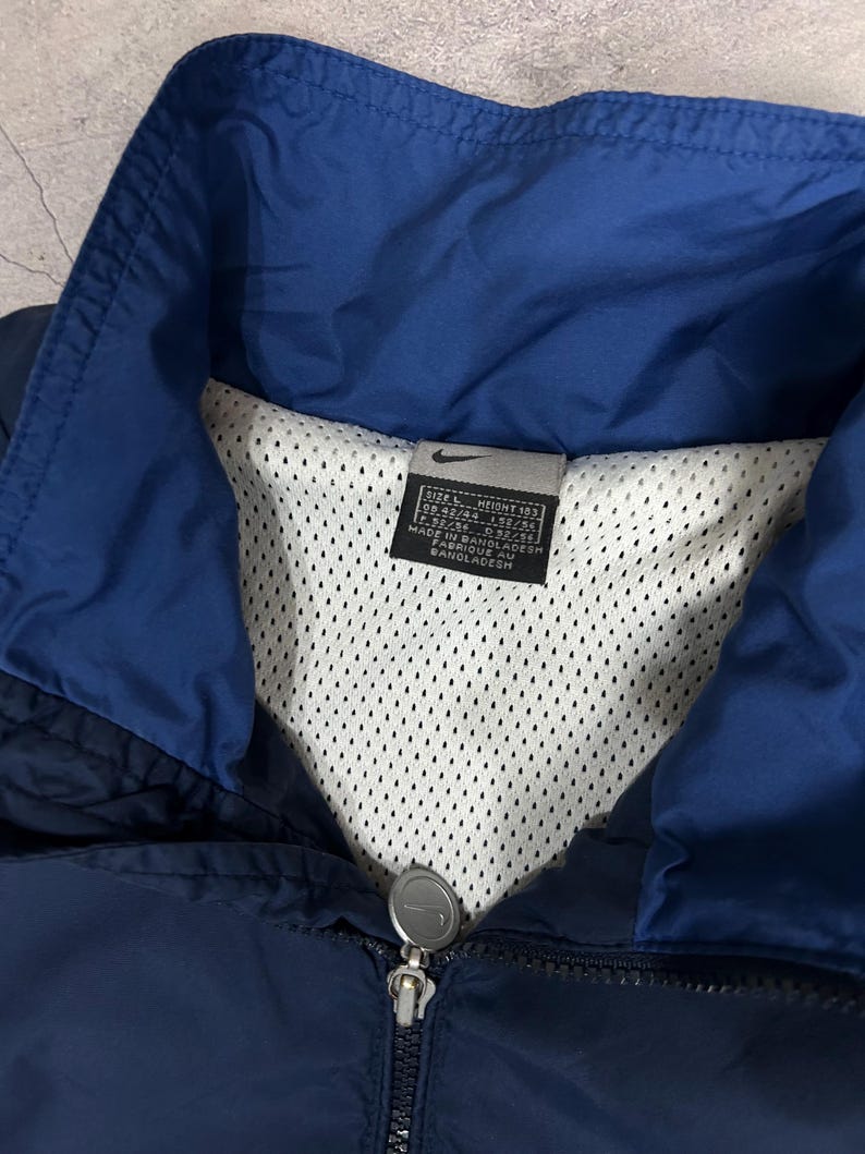 Op de afbeelding: Marineblauwe en koningsblauwe Nike jas met mesh voering. De jas heeft een ritssluiting en een opstaande kraag. Het Nike-logo is zichtbaar op de ritstrekker. Het label geeft maat L aan en is gemaakt in Bangladesh.