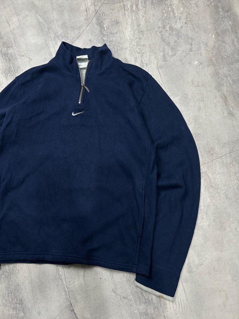 K&ouml;nnte beinhalten: Marineblaues Langarm-Pullover mit Stehkragen und silbernem Rei&szlig;verschluss. Mit kleinem wei&szlig;em Nike-Logo auf der Brust. Die Manschetten und der Kragen haben einen wei&szlig;en Besatz.