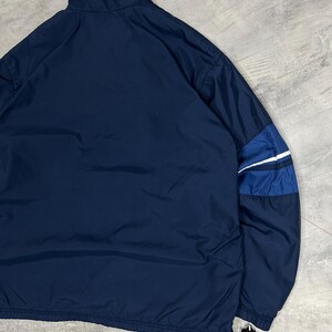 Op de afbeelding: Marineblauwe jas met een hoge kraag en een trekkoord in de zoom. De jas heeft een blauw-wit streepdetail op de mouw. De jas is gemaakt van een lichtgewicht materiaal.