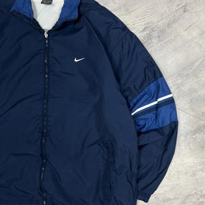 Op de afbeelding: Marineblauwe Nike trainingsjack met een witte mesh voering en een ritssluiting over de hele lengte. De jas heeft een witte Nike swoosh-logo op de borst en blauwe en witte strepen op de rechter mouw. De kraag en manchetten zijn donkerder blauw.