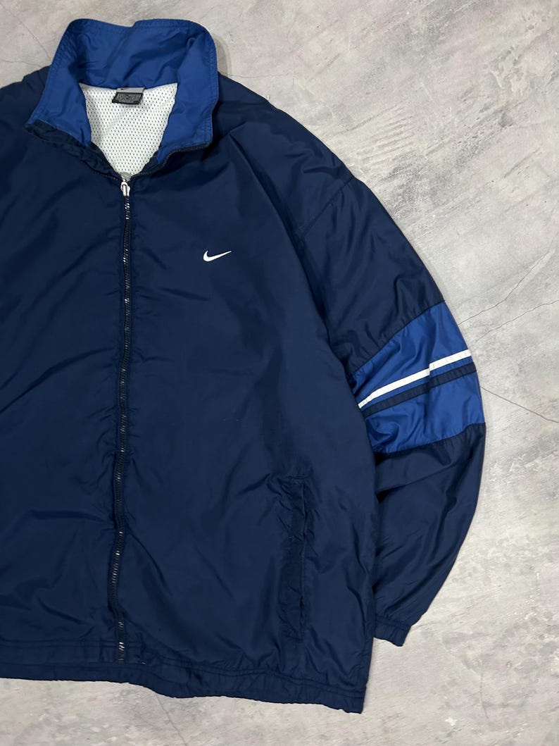 Op de afbeelding: Marineblauwe Nike trainingsjack met een witte mesh voering en een ritssluiting over de hele lengte. De jas heeft een witte Nike swoosh-logo op de borst en blauwe en witte strepen op de rechter mouw. De kraag en manchetten zijn donkerder blauw.
