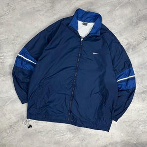 Op de afbeelding: Marineblauwe jas met ritssluiting en wit Nike-logo op de borst. De jas heeft blauwe en witte strepen op de mouwen en een blauwe kraag. De jas heeft een trekkoord aan de onderkant.
