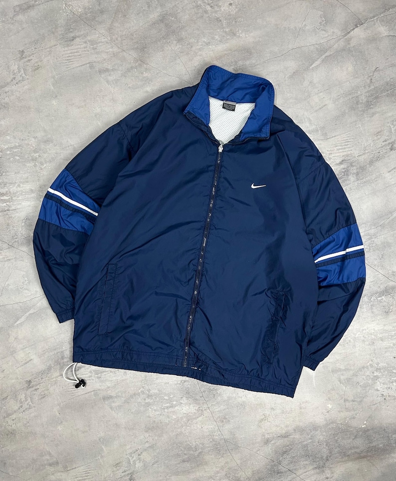 Op de afbeelding: Marineblauwe jas met ritssluiting en wit Nike-logo op de borst. De jas heeft blauwe en witte strepen op de mouwen en een blauwe kraag. De jas heeft een trekkoord aan de onderkant.