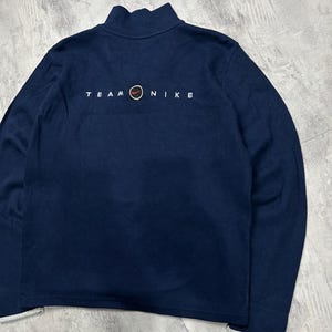 K&ouml;nnte beinhalten: Marineblaues Sweatshirt mit Stehkragen und langen &Auml;rmeln. Der Text "TEAM NIKE" ist in Wei&szlig; auf der R&uuml;ckseite aufgedruckt, mit einem roten und orangefarbenen Kreissymbol. Die Manschetten und der Saum haben einen grauen Besatz.