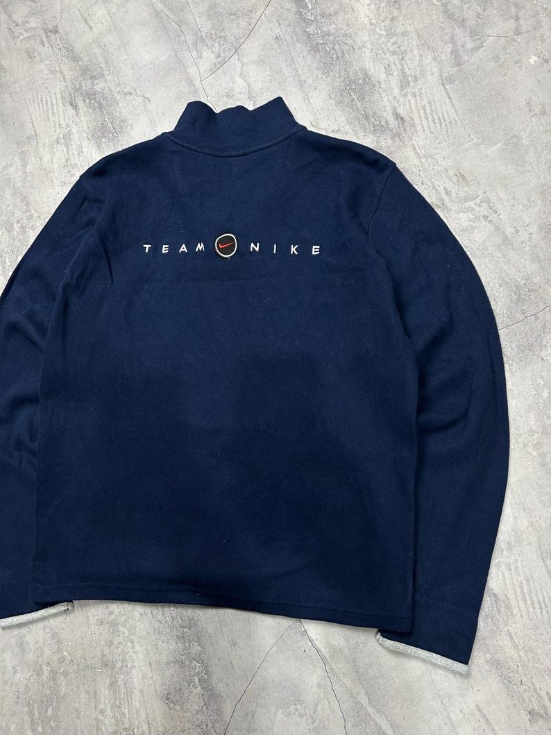 K&ouml;nnte beinhalten: Marineblaues Sweatshirt mit Stehkragen und langen &Auml;rmeln. Der Text "TEAM NIKE" ist in Wei&szlig; auf der R&uuml;ckseite aufgedruckt, mit einem roten und orangefarbenen Kreissymbol. Die Manschetten und der Saum haben einen grauen Besatz.