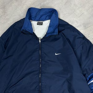 Op de afbeelding: Marineblauwe Nike windjack met een wit Nike swoosh-logo op de borst. De jas heeft een blauwe kraag en een witte mesh voering. De jas is dichtgeritst. Klassieke atletische stijl.