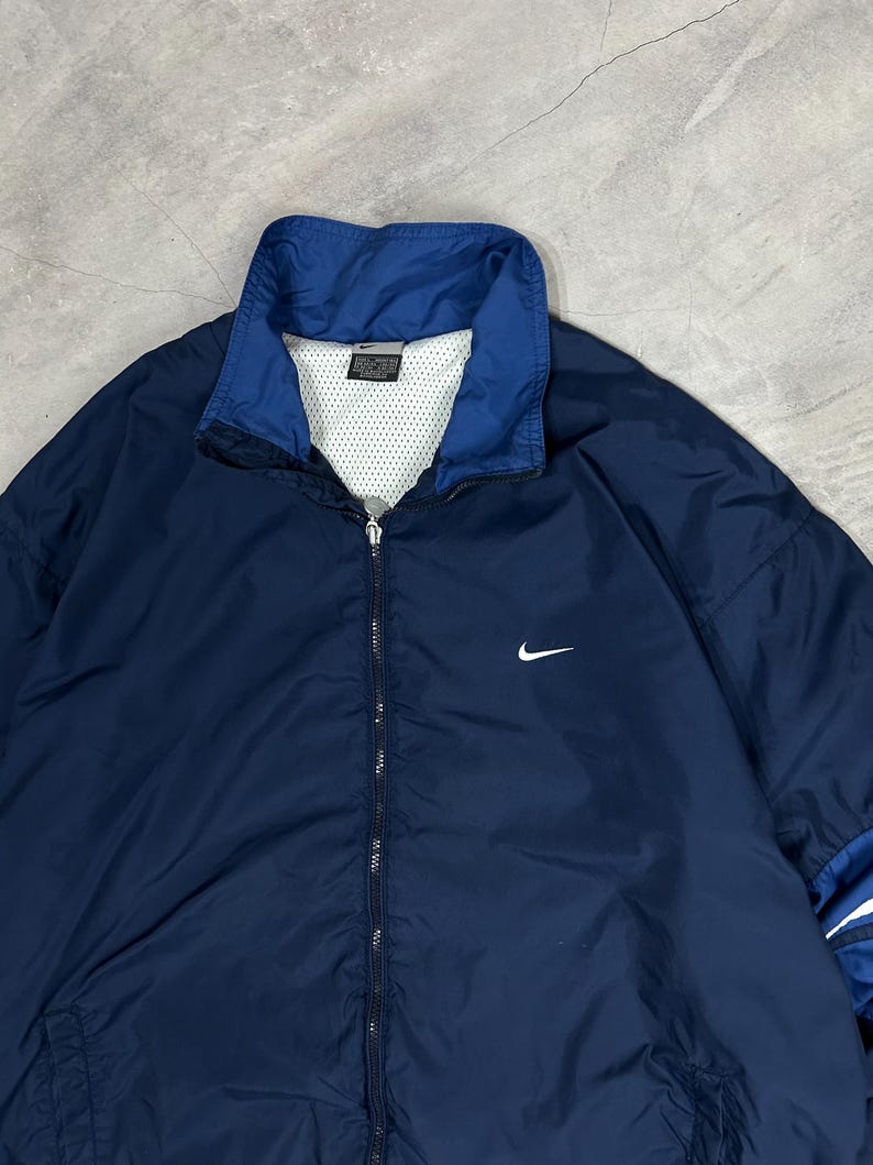 Op de afbeelding: Marineblauwe Nike windjack met een wit Nike swoosh-logo op de borst. De jas heeft een blauwe kraag en een witte mesh voering. De jas is dichtgeritst. Klassieke atletische stijl.