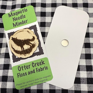 May include: A magnetic needle minder with a wooden wolf design. The minder is attached to a white card with a magnet on the back. The card also has the text "Otter Creek Floss and Fabric".