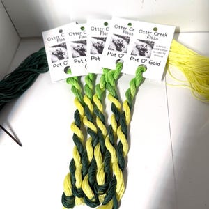 May include: Bundles of green and yellow embroidery floss are displayed, with several skeins of twisted thread. The floss is labeled "Otter Creek Floss" and "Pot O' Gold". The image shows a variety of colors and textures.