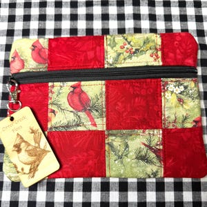May include: A quilted zippered pouch with a cardinal bird design. The pouch is made of red fabric squares and squares with a green and white pattern. A wooden tag with a bird illustration hangs from the zipper.