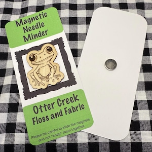 May include: A magnetic needle minder with a wooden frog design. The frog is inside a black frame, with the text "Magnetic Needle Minder" above it and "Otter Creek Floss and Fabric" below. A separate white card with a magnet is also shown.