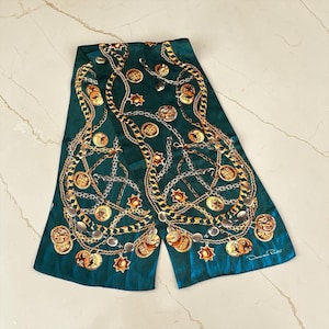 May include: A rectangular emerald green scarf featuring a printed pattern of gold and silver chains, circular medallions, and star-shaped charms. The design includes intricate metallic links and coin-like motifs throughout. The name Oscar de la Renta is printed in white in the bottom right corner.