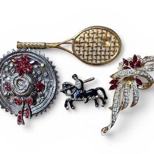 May include: A collection of vintage brooches. Includes a silver-toned circular brooch with red accents, a gold tennis racket brooch, a black equestrian brooch, and a gold bow brooch with red and clear stones.
