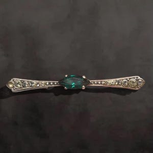 May include: A silver bar pin with a central, emerald-green marquise-cut stone. The pin is accented with smaller, round, clear stones and decorative detailing along its length. The pin is set against a dark background.