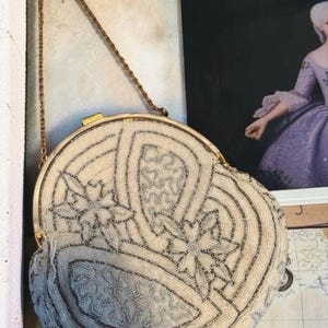 May include: A round, cream-colored beaded handbag featuring intricate silver-toned beadwork in floral and geometric patterns. The bag includes a gold-toned metal frame, a secure clasp closure, and a thin, matching gold-toned metal chain shoulder strap.