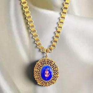 May include: A gold-toned necklace with a detailed chain and a pendant. The pendant features a blue oval center with a floral design and is surrounded by an ornate, openwork frame. The necklace is displayed against a soft, neutral background.