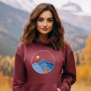 Op de afbeelding: Een bordeauxrode hoodie met een afbeelding van een bergketen met een zon en een kleine oranje boom in een cirkel op de voorkant.