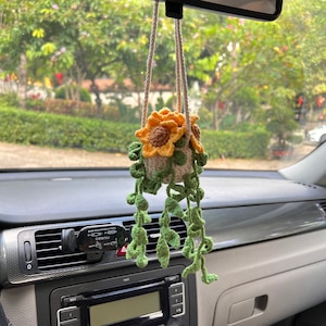 May include: A handmade crochet car charm featuring a sunflower design. The charm includes a beige pot with yellow and brown sunflowers and green vines. The charm hangs from a beige string, adding a touch of nature to the car's interior.