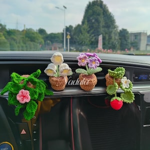 May include: Crocheted car accessories featuring miniature potted plants. The collection includes a green leafy plant with a pink flower, white and yellow flowers, purple flowers, and a strawberry with white flowers. The accessories are handmade and add a touch of nature to the car interior.