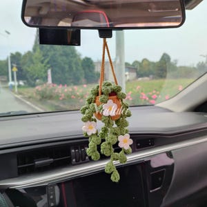 May include: A handmade crochet car charm featuring a hanging plant design. The charm includes a small orange pot with green leaves and white flowers. The word "Vachida" is written on the pot. The charm is suspended from a brown string.