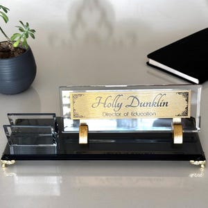 May include: A black and clear glass desk nameplate with gold detailing. The nameplate displays "Holly Dunklin, Director of Education" in gold script. A small potted plant and a black notebook are also present.