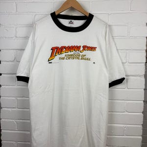 May include: White t-shirt with black trim around the collar and sleeves. The shirt features the text "INDIANA JONES and the KINGDOM OF THE CRYSTAL SKULL" in orange and yellow lettering. The shirt is hanging on a wooden hanger.