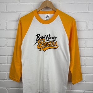 May include: White and gold baseball style t-shirt with the text "Bad News Bears" in black and gold script. The shirt has three-quarter length sleeves and a crew neck. The shirt is hanging on a wooden hanger against a white brick wall.
