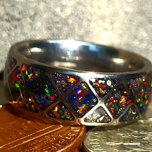 May include: A silver-colored ring with a geometric design filled with colorful, iridescent material. The ring's band is thick and polished, and the inlaid material displays hues of red, blue, green, and yellow. The ring is a piece of jewelry.
