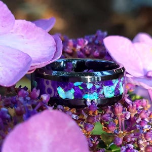 May include: A black tungsten carbide ring with a purple and blue opal inlay. The ring is set against a background of purple flowers.