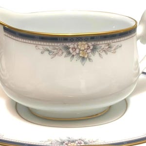 May include: White porcelain gravy boat with a matching saucer. The gravy boat has a gold rim and a floral design with blue accents. The saucer has a matching blue and gold border with floral accents. A classic addition to any dining set.