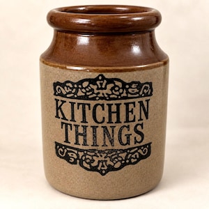 May include: A cylindrical ceramic crock with a glossy brown rim and a speckled beige body. The front features black text that reads KITCHEN THINGS, enclosed within two decorative black filigree borders above and below the lettering.