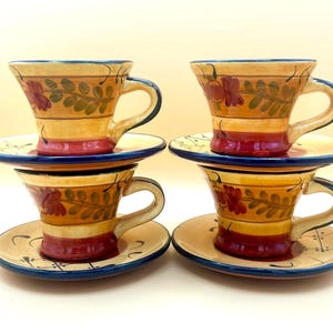 May include: Four ceramic cups and saucers featuring a hand-painted design with yellow, red, and green floral motifs. Each piece has a dark blue rim and base, with a flared cup shape and a matching saucer decorated with thin black lines.