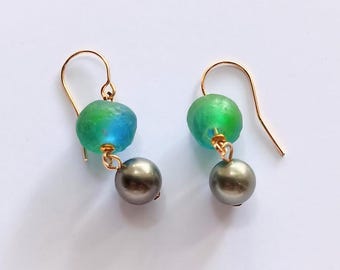 Puede incluir: Un par de pendientes colgantes con ganchos dorados, cada uno con una cuenta redonda, texturizada, translúcida de color verde y azul, combinada con una cuenta esférica metálica de color gris oscuro que cuelga justo debajo.