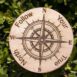 Op de afbeelding: Een houten kompas met de woorden "Follow Your True North" gegraveerd rond de kompasroos. Het kompas ligt op een groene, bladerrijke achtergrond.