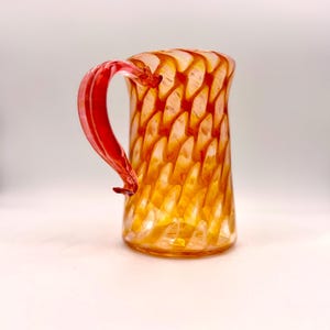May include: A decorative glass pitcher with a red handle. The pitcher has a pattern of orange and yellow swirls. The handle is a solid red color. The pitcher is sitting on a white surface.