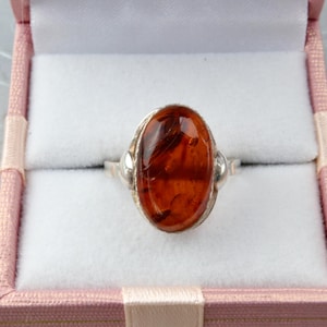 Puede incluir: Un anillo de plata con una piedra de &aacute;mbar de forma ovalada. El anillo est&aacute; en una caja de anillo rosa.