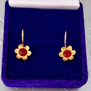 Puede incluir: Un par de pendientes dorados con un diseño en forma de flor y una piedra circular roja en el centro. Los pendientes tienen ganchos curvos y se presentan dentro de un joyero rectangular de terciopelo azul con un forro interior blanco.