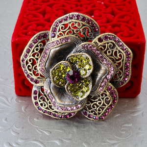 Op de afbeelding: Een bloemvormige broche met filigraan bloemblaadjes van antiek bronskleurig metaal. De buitenste blaadjes zijn ingelegd met roze kristallen, terwijl de binnenste blaadjes zijn gevuld met limoengroene kristallen en een paars kristal in het midden.