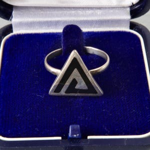 May include: A silver-toned ring featuring a triangular face with a black geometric inlay. The ring is displayed inside a dark blue, square-shaped presentation box with a plush, deep blue velvet interior lining and a white satin trim.