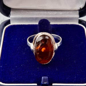 Puede incluir: Un anillo de plata con una piedra de &aacute;mbar de forma ovalada engastada en un engaste de plata. El anillo est&aacute; en una caja de anillo de terciopelo azul.